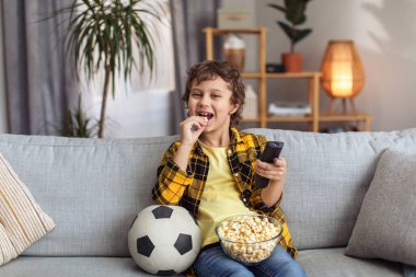 TV entertainment. Happy little boy spending weekend at home, watching sport match and eating popcorn, resting on sofa in evening, free space