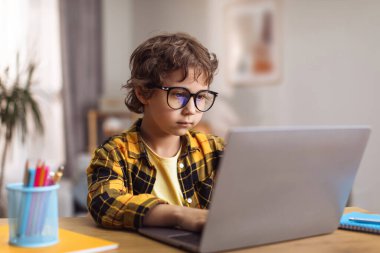 Smart generation. Concentrated little boy wearing eyeglasses typing on laptop at home, creating new game online, sitting at desk, free space