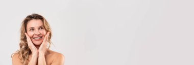 Beauty Ad. Smiling beautiful middle aged woman touching cheeks and looking away, wide horizontal banner with attractive female posing over grey studio background, panorama with copy space
