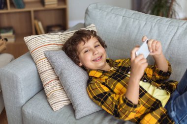 Modern kids leisure. Cute little boy playing games on mobile phone, resting on sofa at home, lying and smiling to camera, empty space