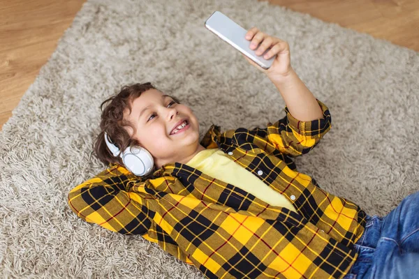 Social media content. Positive little boy wearing wireless headphones making selfie on smartphone, smiling to device, lying on floor at home, free space