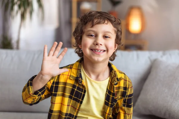 Hello. Camera pov portrait of positive little boy waving hand, video calling or recording blog at home interior, sitting on sofa