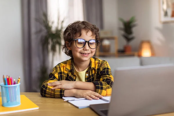 Happy learning. Clever little boy wearing eyeglasses watching lesson online on laptop, sitting at home workplace and smiling, free space