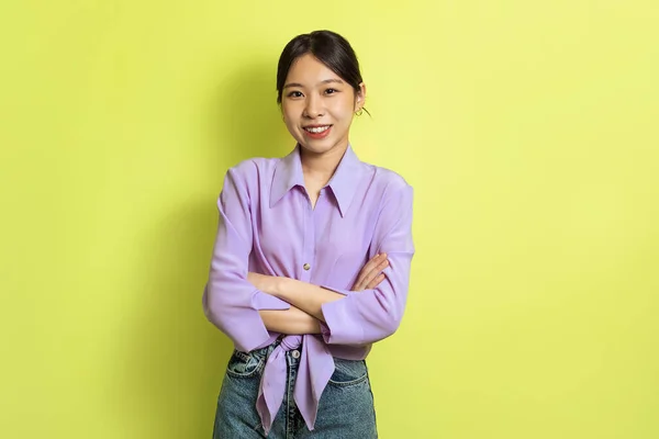 Cheerful Asian Millennial Lady Crossing Hands Posing Smiling Looking At Camera Standing Over Yellow Background In Studio. Female Beauty, Fashion And Self Confidence Concept