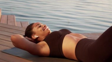 Abs workout. Close up portrait of happy active african american woman in earphones practicing crunches, exercising on wooden pier near lake, slow motion, free space
