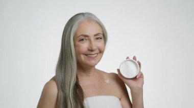 Anti aging body cosmetics. Pretty senior grey haired lady wrapped in towel showing jar with pampering cream, smiling to camera with creamy heart on shoulder, white studio background, slow motion