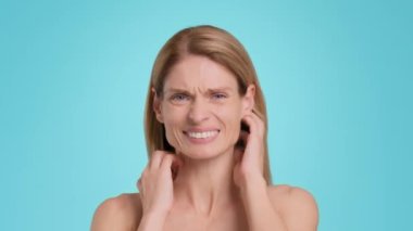 Allergy symptoms. Upset middle aged woman having rash, scratching her neck and head, frowning face, suffering from allergy attack, blue studio background, slow motion