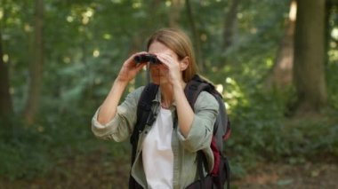 Middle aged active woman tourist peering into distance with binoculars, hiking with backpack in forest, looking for route, slow motion, empty space