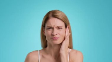 Acute dental pain. Close up portrait of unhappy middle aged woman suffering from acute toothache, touching her painful jaw, blue studio background, slow motion