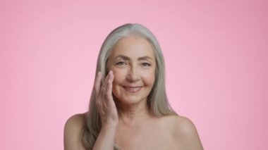 Beauty care cosmetics. Close up portrait of senior grey haired lady rubbing anti aging cream on her face skin, smiling to camera, pink studio background, slow motion