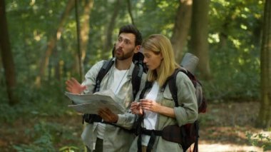 Navigation in woodland. Couple of active middle aged tourists with backpacks using paper map to find right route, discussing way during hiking trip in forest, slow motion