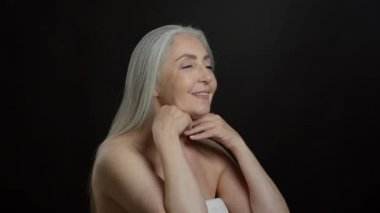 Skin lifting beauty procedures. Positive senior grey haired lady doing self massage, exercising against double chin and skin sagging, black studio background, semi profile, slow motion, free space