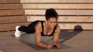Close up portrait of happy sporty african american lady athlete standing in plank position on wooden pier, slow motion, tracking shot, free space