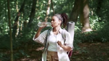 Active weekend. Young happy african american lady walking in forest in summer wood, drinking water and enjoying water, slow motion, tracking shot