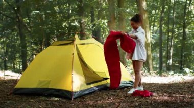 Accommodation arrangement. Young positive african american lady putting sleeping bag into tent, preparing for overnight stay in forest, slow motion