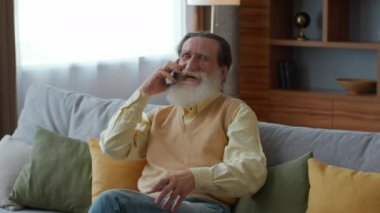 Lovely family relationships. Portrait of happy senior bearded gentleman talking with relatives via cellphone, sitting at home, tracking shot, slow motion, empty space