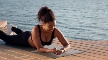 Body strength concept. Young active african american woman stop practicing plank position on pier near water, checking intensivity on smartwatch, slow motion, empty space