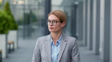 Confident middle aged business lady wearing eyeglasses walking outdoors near modern office building, follow shot, slow motion