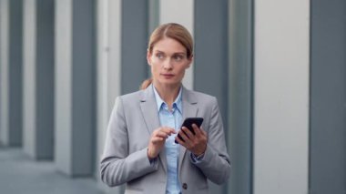 Business and mobile communication. Confident middle aged woman ceo typing on cellphone, chatting with partners, standing outdoor near office building, slow motion