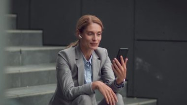 Pleasure communication on distance. Happy middle aged business woman in earbuds video chatting with friends online, resting on office staircase, slow motion, free space