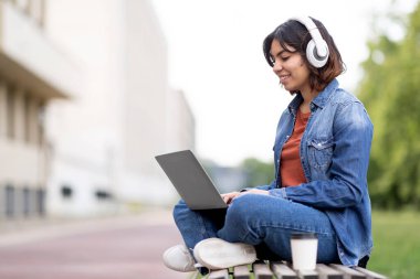 Smiling arab female student in wireless headphones study with laptop outdoors, young middle eastern woman sitting with computer on bench at campus, enjoying distance learning, copy space
