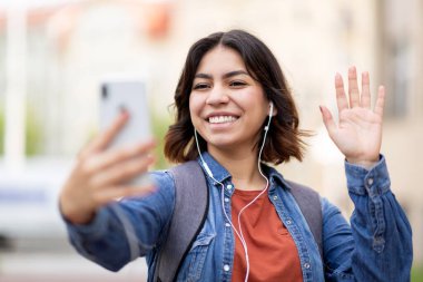 Happy young arab woman making video call via cellphone while standing outdoors, cheerful middle eastern female student wearing earphones waving hand at camera, gesturing hello, communicating online