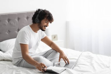 Happy curly bearded young eastern guy studying in bed, using laptop and wireless headset, watching course on Internet or attending webinar, taking notes, wearing pajamas, copy space, side view