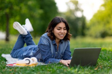 Smiling arab female student using laptop while lying on lawn outdoors, happy young middle eastern woman relaxing in city park with computer, preparing for exams outside, enjoying study online
