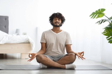 Peaceful millennial indian man in homewear practicing yoga at home, sitting on fitness mat with closed eyes, meditating, copy space, enjoying morning workout, full length shot. Stress relief concept