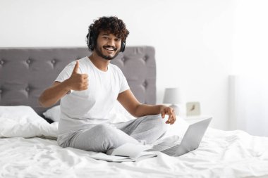 Positive curly millennial eastern guy sitting on bed, using wireless headset and laptop, showing thumb up and smiling, attending webinar on Internet, bedroom interior, copy space