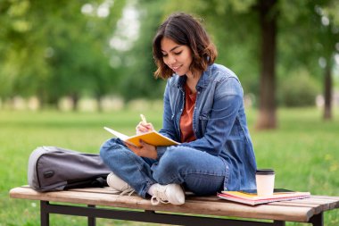 Beautiful arab female student writing in notebook while sitting on bench in park, portrait of smiling young middle eastern woman taking notes to notepad while study outdoors at college campus