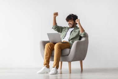 Happy young indian man gabmling online, raising hands up, celebrating victory, sitting in armchair and using laptop over white background, copy space. Gaming, betting on Internet concept