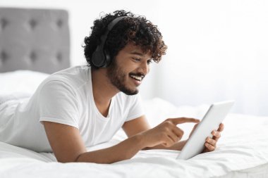 Relaxed young curly hindu guy using modern digital tablet and wireless headphones while chilling in bed at home, watching videos on Internet, playing online video game, copy space, closeup