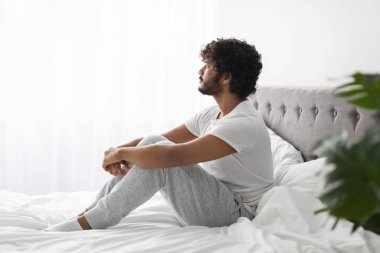 Pensive curly bearded young indian guy in pajamas sitting on bed with legs up at home, hugging his laps, thinking about something, suffering from loneliness, looking at copy space, side view