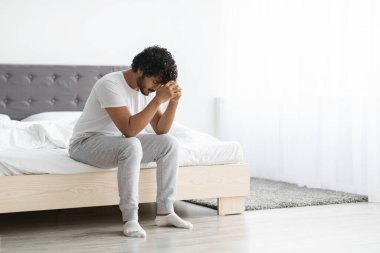Full length shot of depressed curly bearded young indian guy in pajamas sitting on bed alone at home, touching his head, thinking about something, suffering from loneliness, side view, copy space