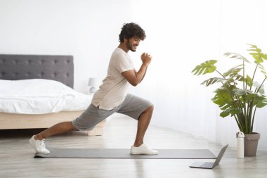 Athletic young indian guy in sportswear exercising at home, working on legs strength, looking at laptop screen and doing workout, enjoying online fitness video, copy space, bedroom interior, panorama