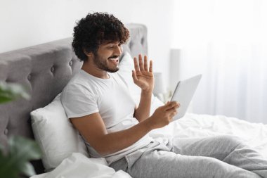 Happy millennial indian guy sitting on bed at home, holding modern digital tablet, waving at gadget screen and smiling, having video call with friend or lover, side view, copy space