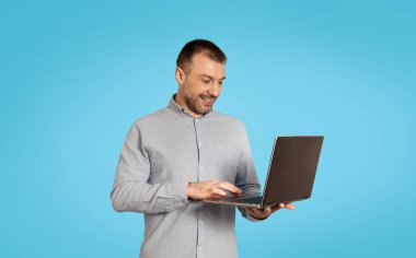Happy Middle Aged Male Using Laptop Computer Websurfing And Working Online Standing Over Blue Studio Background. Internet Technology And Freelance Concept