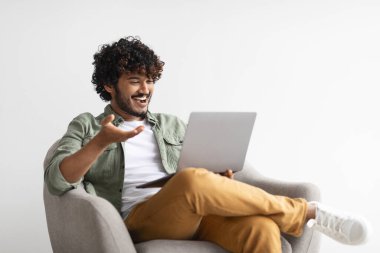 Relaxed handsome millennial eastern guy having video call with friend, sitting in armchair over white background, using computer, looking at laptop screen, smiling and gesturing, copy space