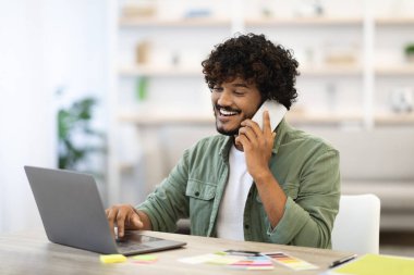 Cheerful handsome young indian guy with beard and curly hair entrepreneur working online, sitting at workdesk at home, using laptop and having phone conversation with business partner, copy space