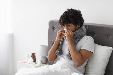 Sick millennial eastern guy with scarf around his neck staying in bed at home, using tissue, sneezing nose, curly young man suffering from coronavirus, copy space. Flu, COVID-19 concept