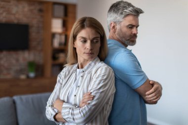 Offended upset adult european husband ignores wife, man and woman stand back to back in room interior. Relationship problems at home, conflict, stress, negative emotions and quarrel, disagreement