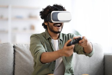 Modern technologies and domestic entertainment concept. Young dark-skinned curly man trying virtual reality at home, using headset VR glasses and holding joystick, playing video games, copy space