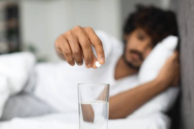 Unhappy millennial indian guy suffering from headache, waking up in morning, lying in bed, putting pills into glass of water, wearing pajamas, blurred background. Hangover, migraine concept