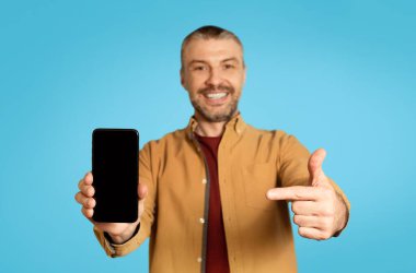 Happy Middle Aged Male Showing Smartphone Screen To Camera, Pointing Finger Recommending App Over Blue Studio Background. Check This Mobile Offer. Mockup, Selective Focus On Cellphone