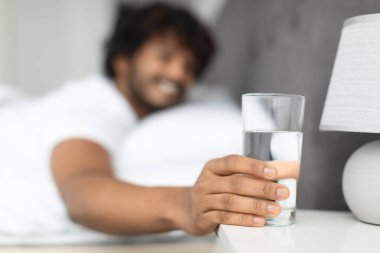 Selective focus on glass of fresh spring water next to eastern guy bed, unrecognizable indian man smiling and taking glass of water after waking up in the morning, copy space. Hydration concept