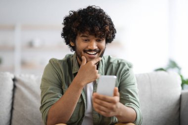 Cheerful curly millennial hindu guy in casual sitting on couch at home, using brand new cell phone and smiling, young eastern man checking newest mobile app, copy space, closeup shot