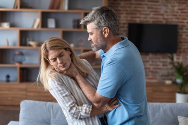 Aggressive angry european husband strangles behind proud frightened afraid wife in living room interior. Beating, conflict at home, pressure, domestic violence, abuse in married couple and fighting