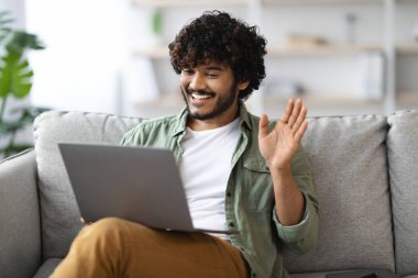 Positive handsome millennial eastern guy having video call with friend, sitting on couch at home, using modern computer, looking at laptop screen, smiling and gesturing, copy space
