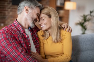 Cheerful adult european man and woman hugging and enjoying tender moment and spare time, sitting together on sofa in living room interior. Love, romantic, family relationships, people emotions at home